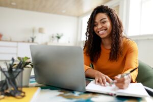 A student writing in a notebook and smiling at her laptop.