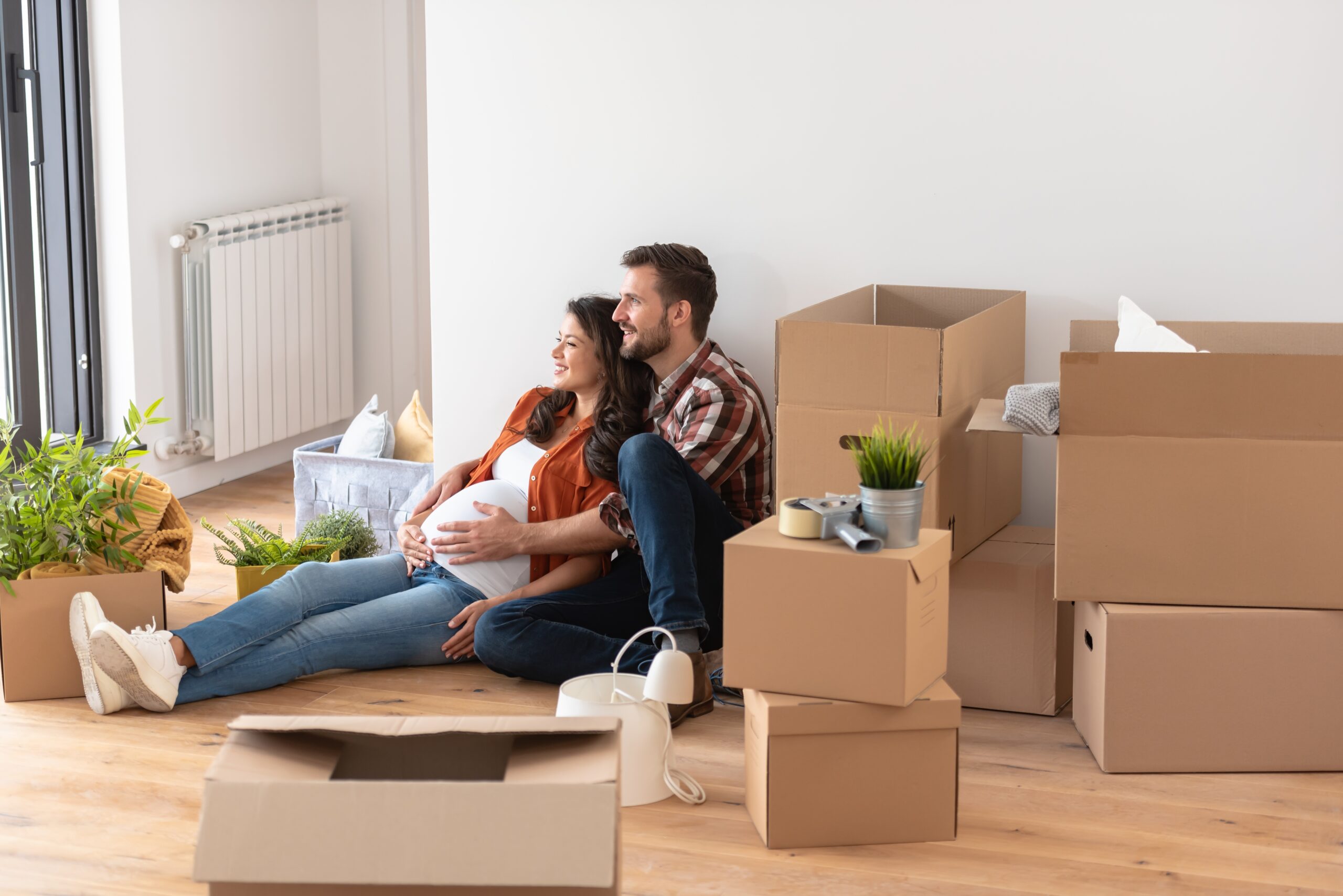 A couple with a pregnant woman sat on the floor with a pile of boxes around them.