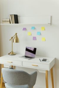 A home office setup with a white desk with laptop on it and a lamp and a white shelf above the desk with a grey chair and Post-it notes stuck on the wall.
