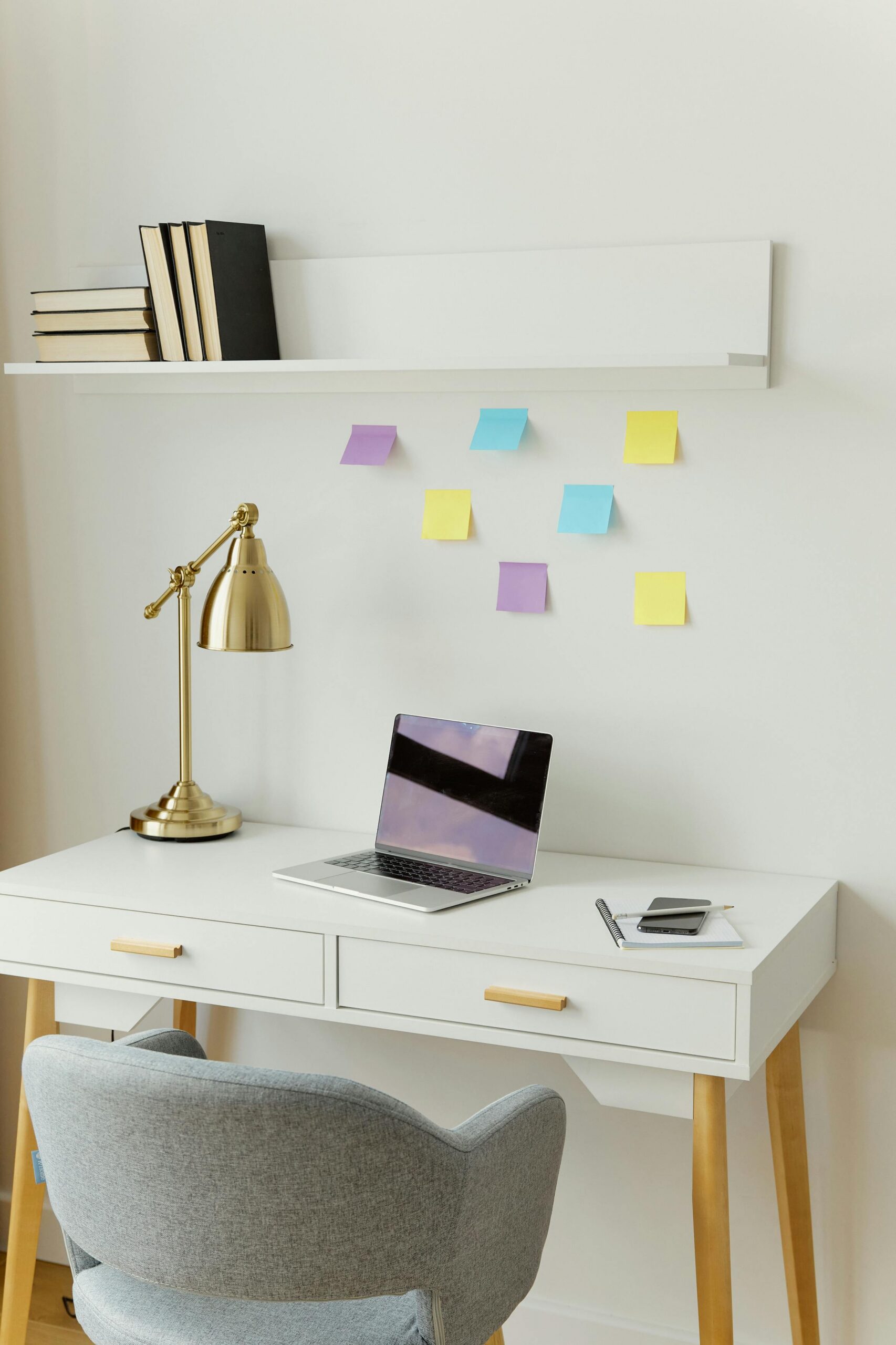 A home office setup with a white desk with laptop on it and a lamp and a white shelf above the desk with a grey chair and Post-it notes stuck on the wall.